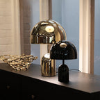 Beside a smaller matching lamp in black, this Clérachic table lamp in gold sits on a dark shelf near a window.