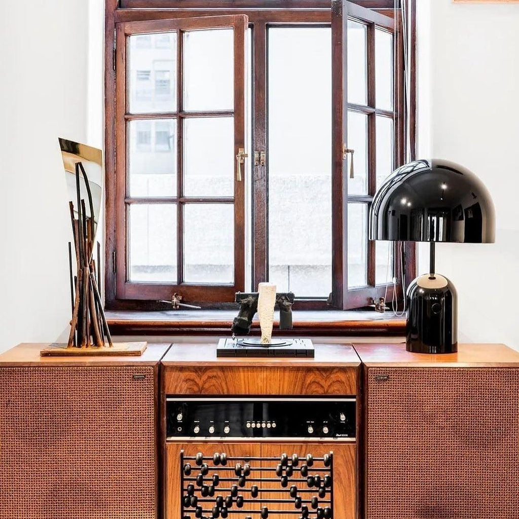 This Clérachic black table lamp sits on a vintage wooden stereo cabinet by a window, beside dried branches in a vase.