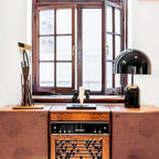 This Clérachic black table lamp sits on a vintage wooden stereo cabinet by a window, beside dried branches in a vase.
