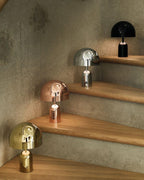Four finishes of this Clérachic table lamp, black, chrome, rose gold and gold, are arranged along wooden stair steps.