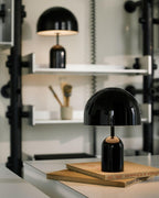 This Clérachic black table lamp stands on a white shelving unit beside small pots and stacked books.