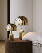 Three warm-toned finishes of this Clérachic table lamp sit together on a round cork pouf beside a white-linen bed.