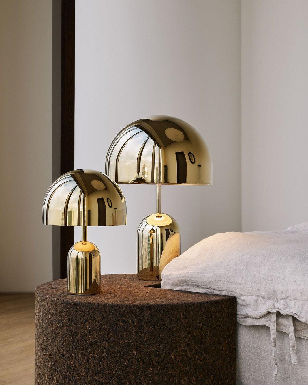 Three warm-toned finishes of this Clérachic table lamp sit together on a round cork pouf beside a white-linen bed.