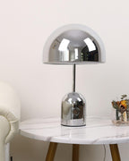 On a round white marble table beside a white leather chair, this Clérachic chrome table lamp sits near cut flowers.