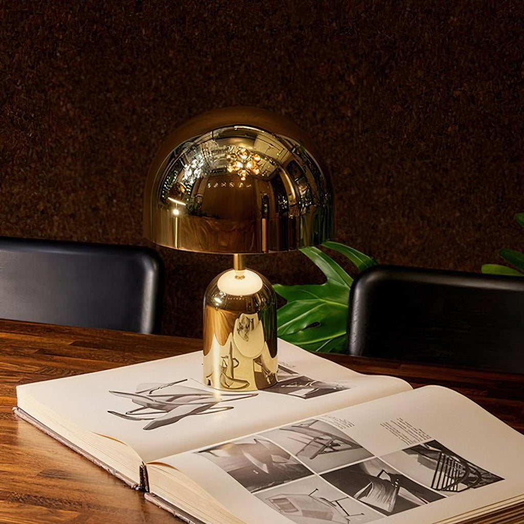 This Clérachic table lamp in gold stands on an open book atop a wood table, with a dark leafy backdrop.