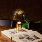 This Clérachic table lamp in gold stands on an open book atop a wood table, with a dark leafy backdrop.