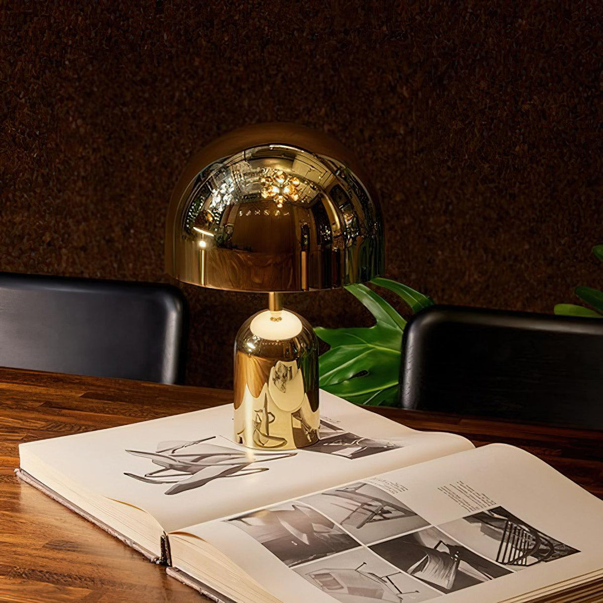 This Clérachic table lamp in gold stands on an open book atop a wood table, with a dark leafy backdrop.