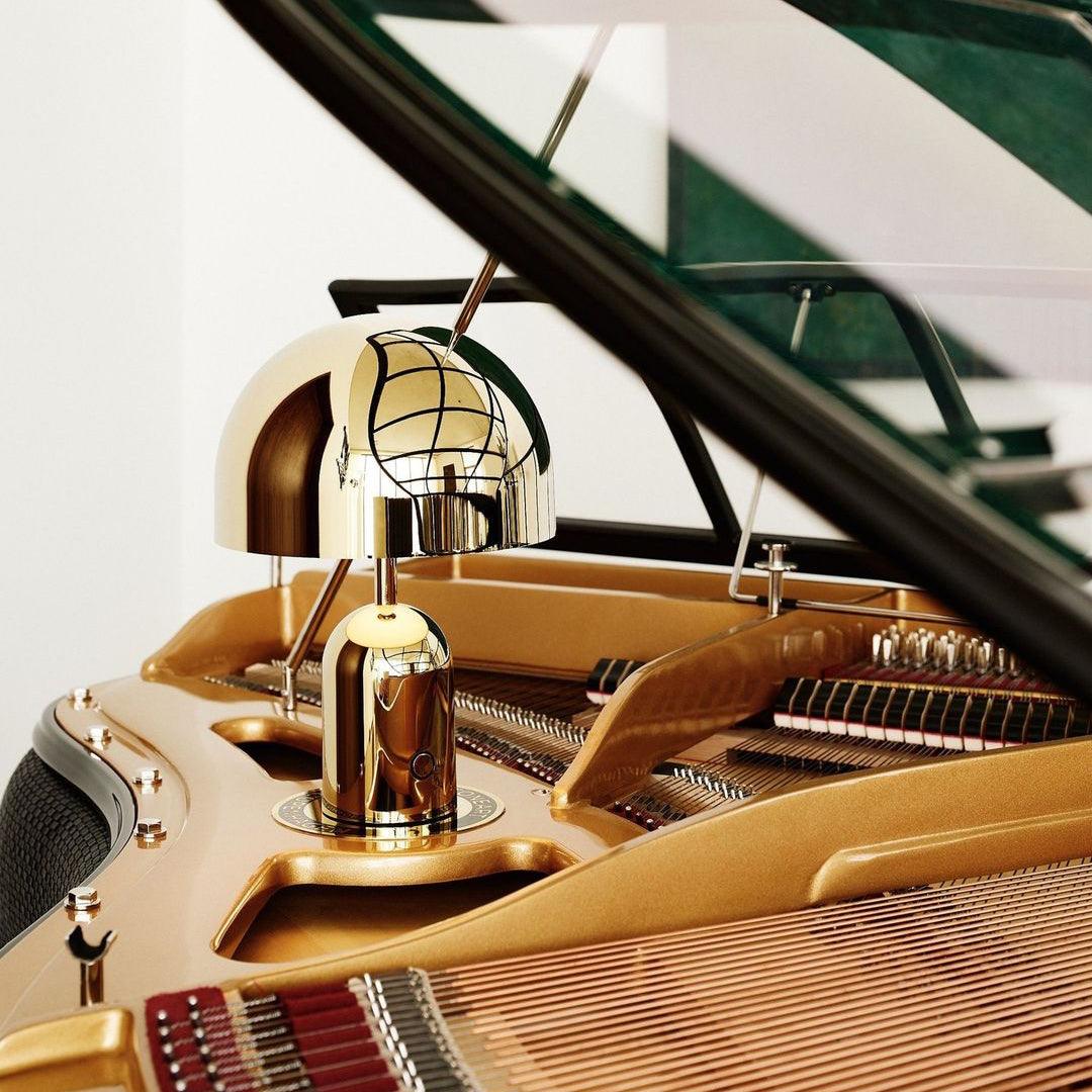 Beside an articulated piano lamp, this Clérachic gold table lamp rests on the open lid of a grand piano.