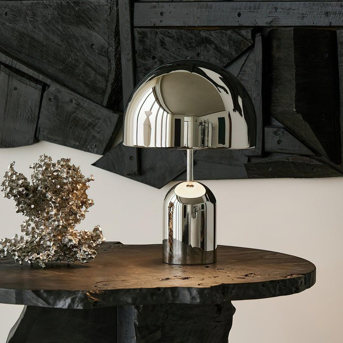 The chrome version of this Clérachic table lamp sits on a round dark-wood table beside a metallic branch ornament.