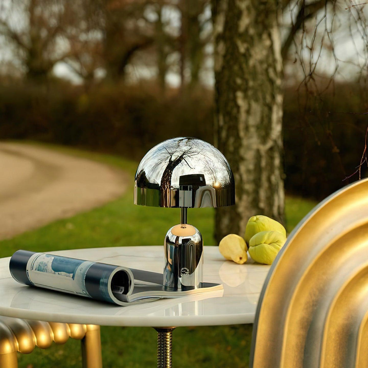 Outdoors on a white table, this Clérachic chrome table lamp reflects the surrounding trees beside a stack of lemons.