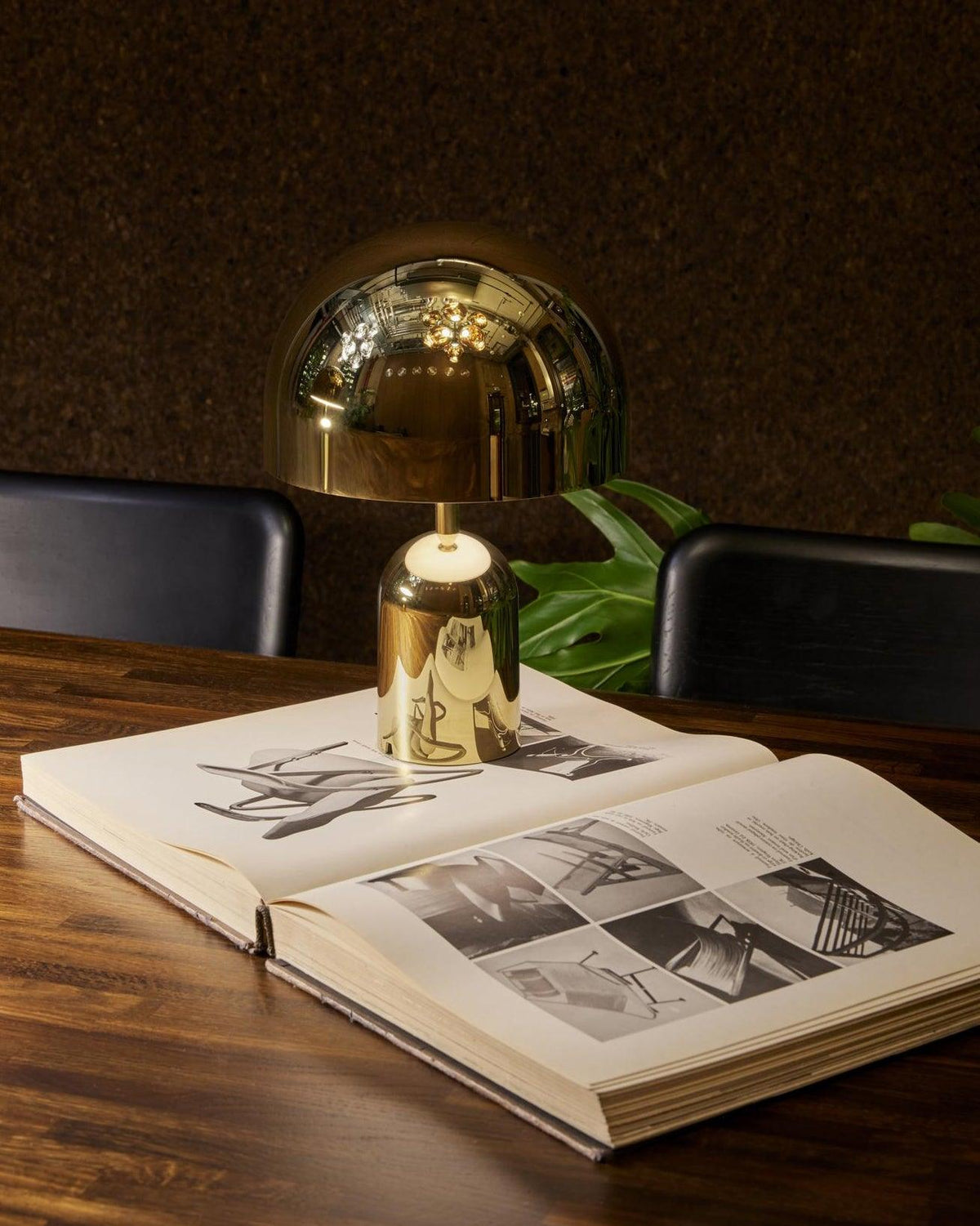 This Clérachic gold table lamp rests on an open book atop a wood table, with dark chairs in the background.