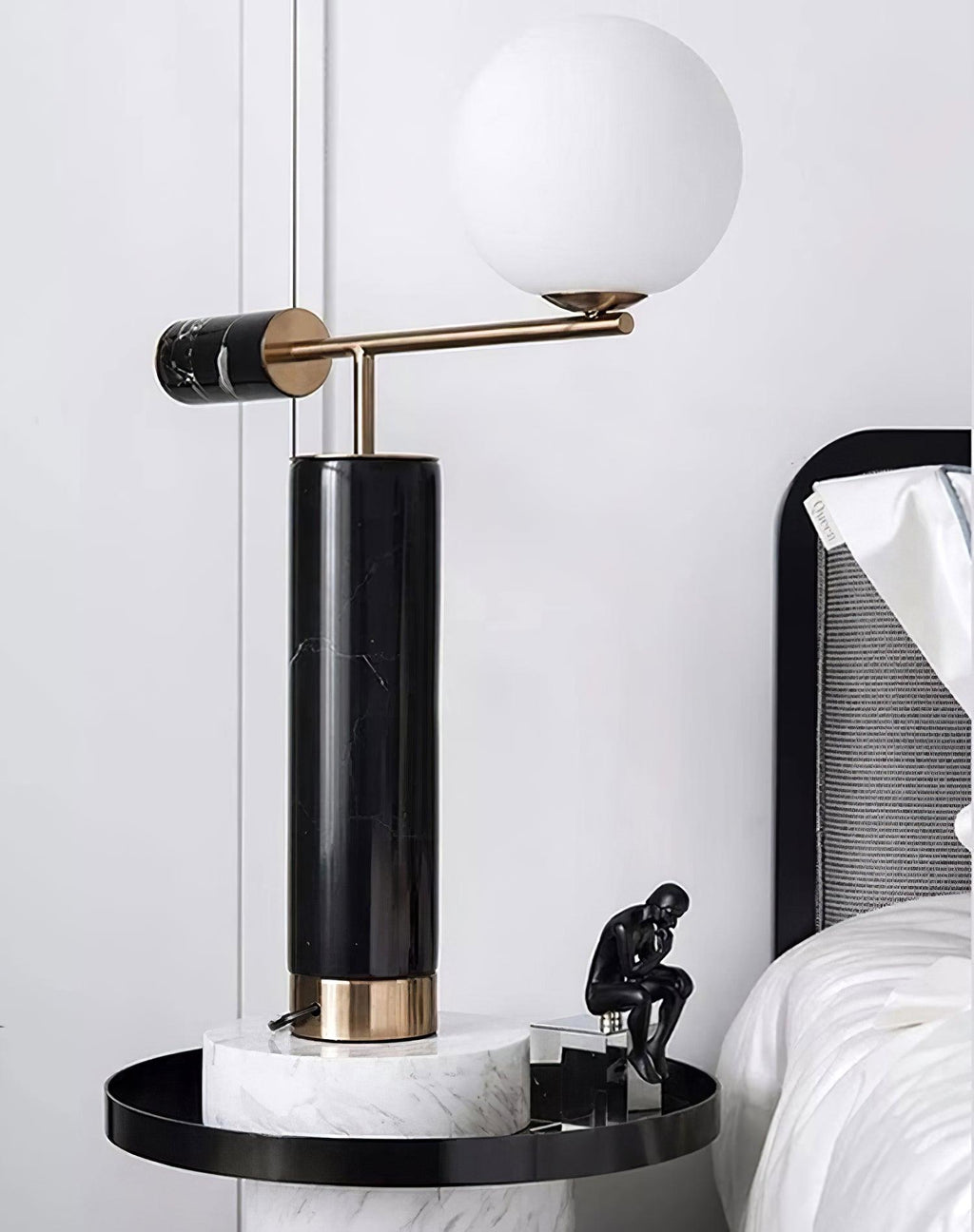 A Clérachic desk lamp glows on a round white marble side table beside a small black sculpture and white bedding.