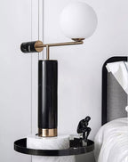 A Clérachic desk lamp glows on a round white marble side table beside a small black sculpture and white bedding.