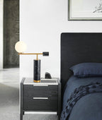 A Clérachic desk lamp glows on a grey nightstand beside a dark upholstered headboard and a phone left charging.