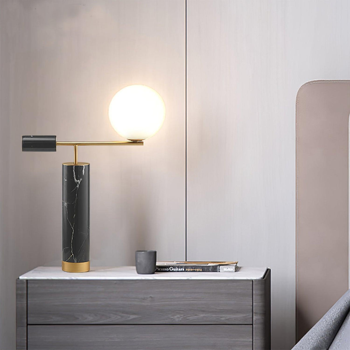 A Clérachic desk lamp glows on a grey dresser beside a mug and folded magazine, with a blush wall behind it.