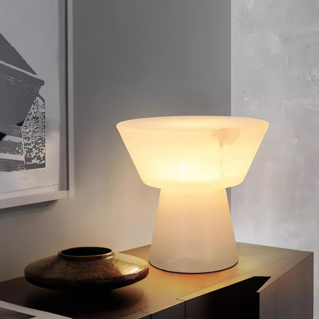 Clérachic alabaster table lamp glowing on a low wooden sideboard beside a bronze bowl and framed artwork on a grey wall.