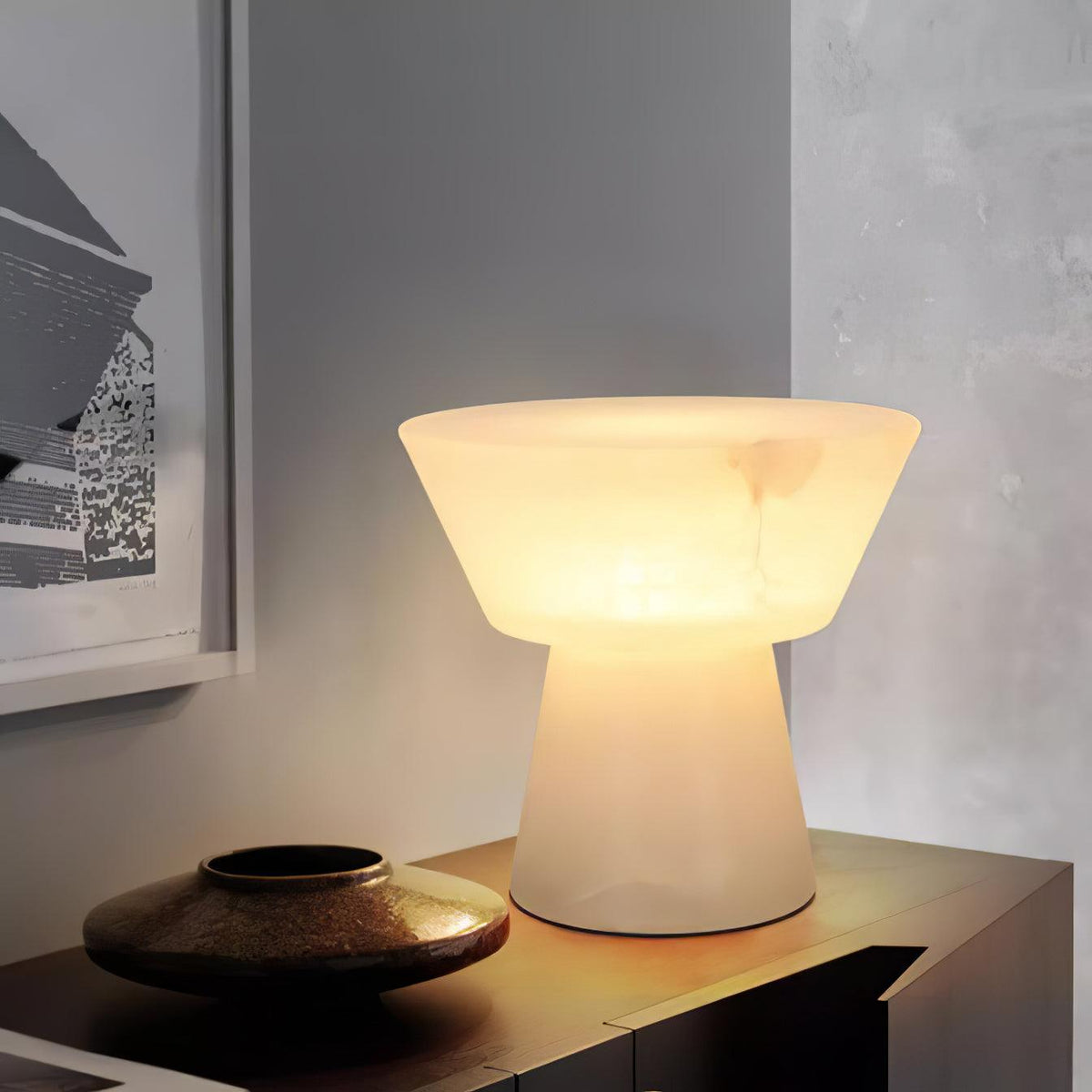 Clérachic alabaster table lamp glowing on a low wooden sideboard beside a bronze bowl and framed artwork on a grey wall.