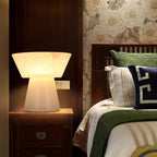 Clérachic alabaster table lamp on a wooden nightstand beside a bed with a green pillow and floral wallpaper behind it.