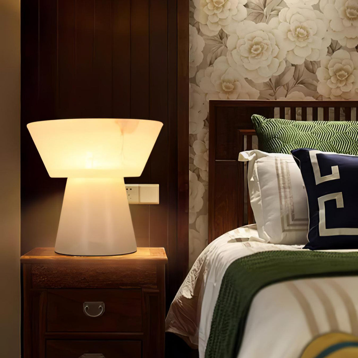Clérachic alabaster table lamp on a wooden nightstand beside a bed with a green pillow and floral wallpaper behind it.