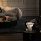 In a moody lounge, the Clérachic alabaster lamp sits lit on a dark round table near a low bench and sofa.