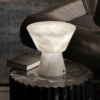 Clérachic hourglass-shaped alabaster table lamp lit on a dark marble side table beside a black leather sofa arm and books.