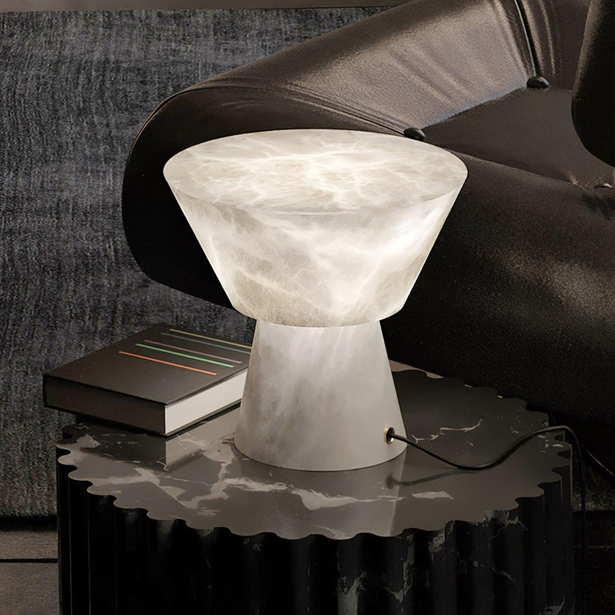 Clérachic hourglass-shaped alabaster table lamp lit on a dark marble side table beside a black leather sofa arm and books.