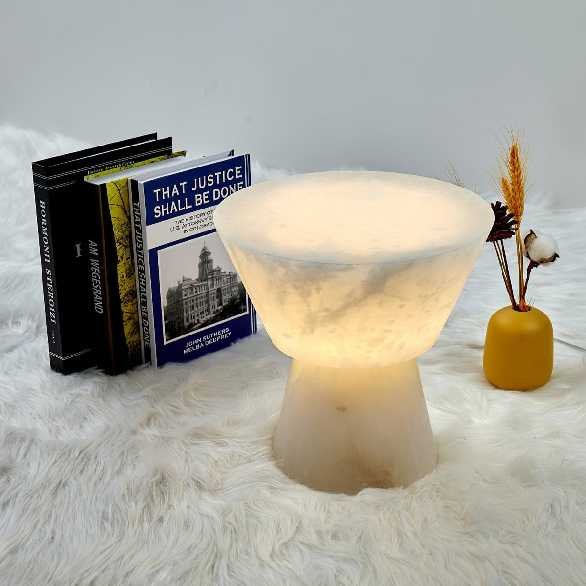 Clérachic alabaster table lamp styled on a white sheepskin rug with stacked books and a mustard vase of dried flowers.
