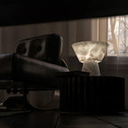 Clérachic alabaster table lamp lit beside a black leather armchair, sheer curtains softening the window light behind it.