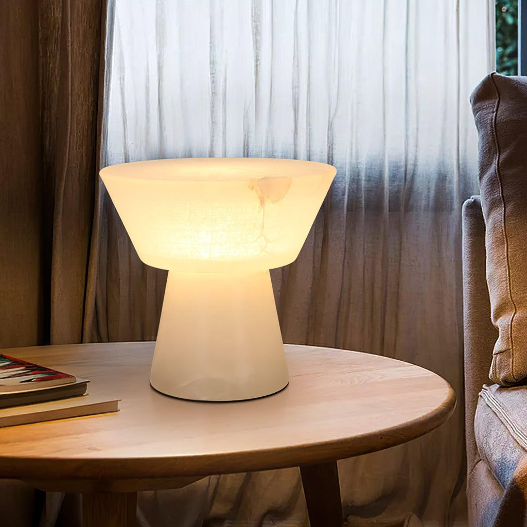 Clérachic alabaster table lamp lit on a round wooden table by sheer curtains, magazines stacked on the table beside it.