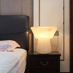 Clérachic alabaster table lamp on a dark wood nightstand beside a bed with a pink pillow and black tufted headboard.