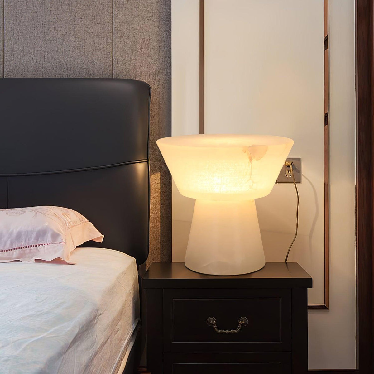 Clérachic alabaster table lamp on a dark wood nightstand beside a bed with a pink pillow and black tufted headboard.