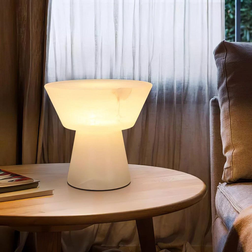 Clérachic alabaster table lamp lit on a round wooden table beside sheer brown curtains and a stack of books.
