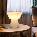 Clérachic alabaster table lamp lit on a round wooden table beside sheer brown curtains and a stack of books.
