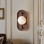 Walnut oval wall light with a lit alabaster disc, mounted on a cream panelled wall beside a framed picture.