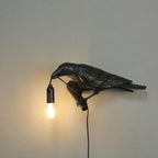 Mounted on a plain wall, this black resin Clérachic bird lamp lowers its beak to the cord, with the bulb glowing warm below.