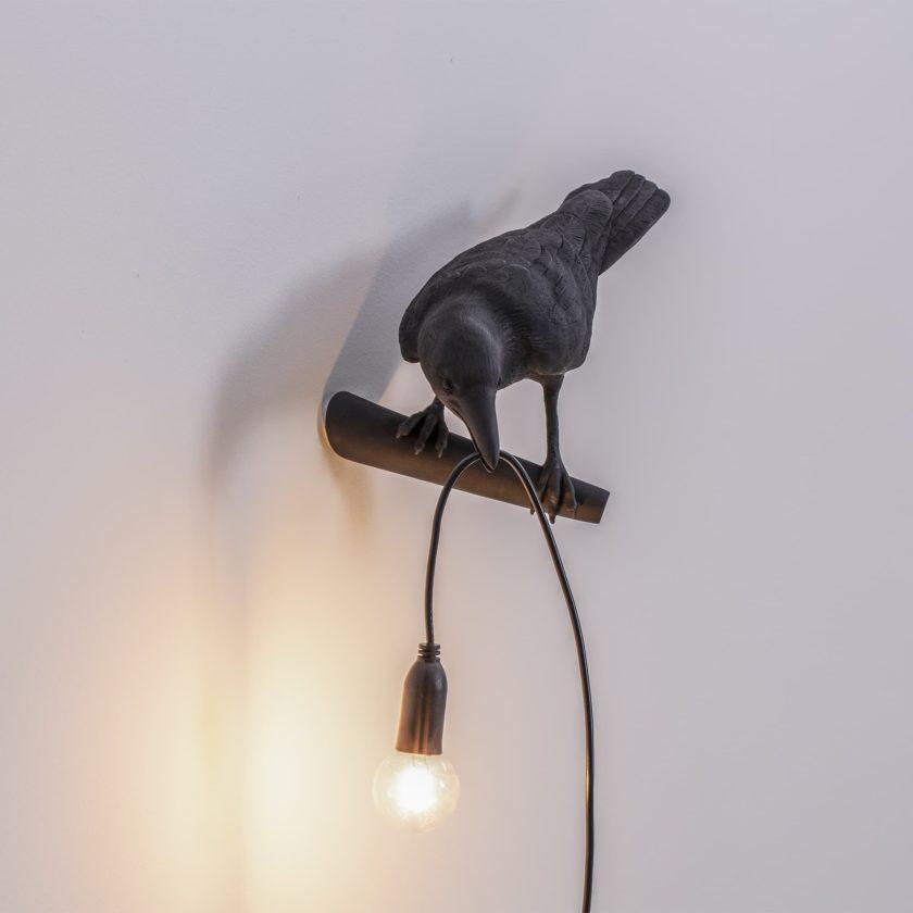 Perched on a wooden dowel bracket, this black Clérachic bird lamp shows its wall mount clearly, bulb lit warm.