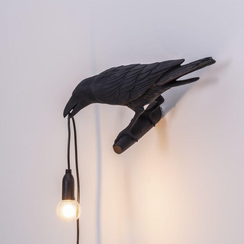 Against a plain grey ground, this black Clérachic bird lamp loops its cord beneath a warmly glowing bulb.