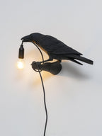 This black bird lamp perches on its wall mount, cord looped below the beak, bulb lit warm against a plain wall, from Clérachic.