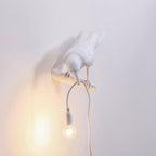 This white resin bird perches on a wall bracket, its warm bulb glowing beneath a lavender-grey backdrop, from Clérachic.