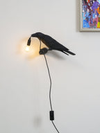 High in a room corner, a black Clérachic bird lamp dangles its lit bulb near the floor, a colourful abstract print above.