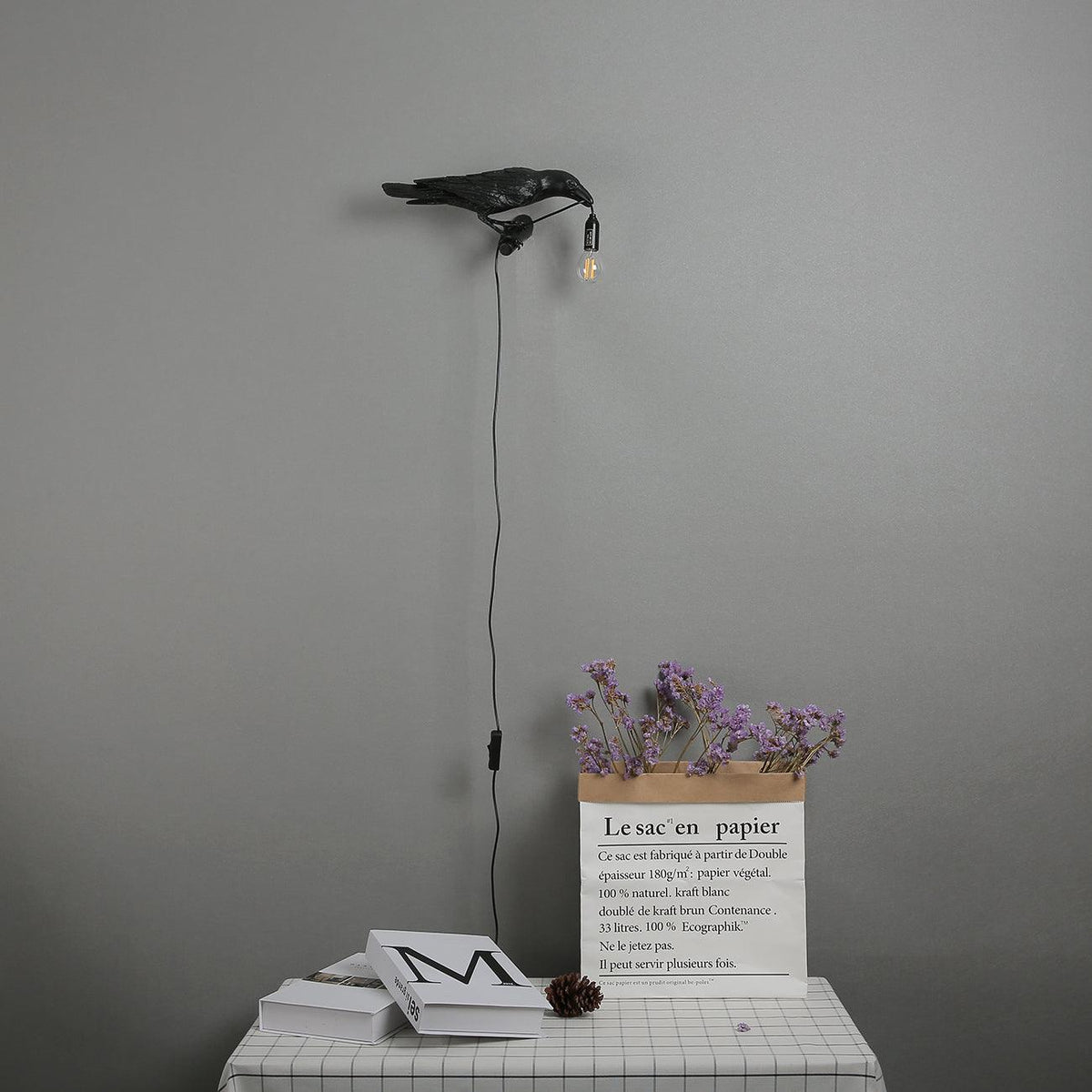 Above a bedside table with dried flowers and stacked books, this black Clérachic bird lamp casts a small warm glow.