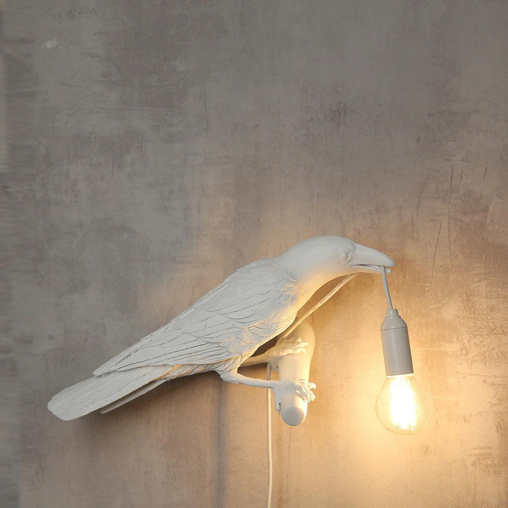 Against a moody, weathered concrete wall, a white Clérachic bird lamp casts warm light from the bulb at its beak.