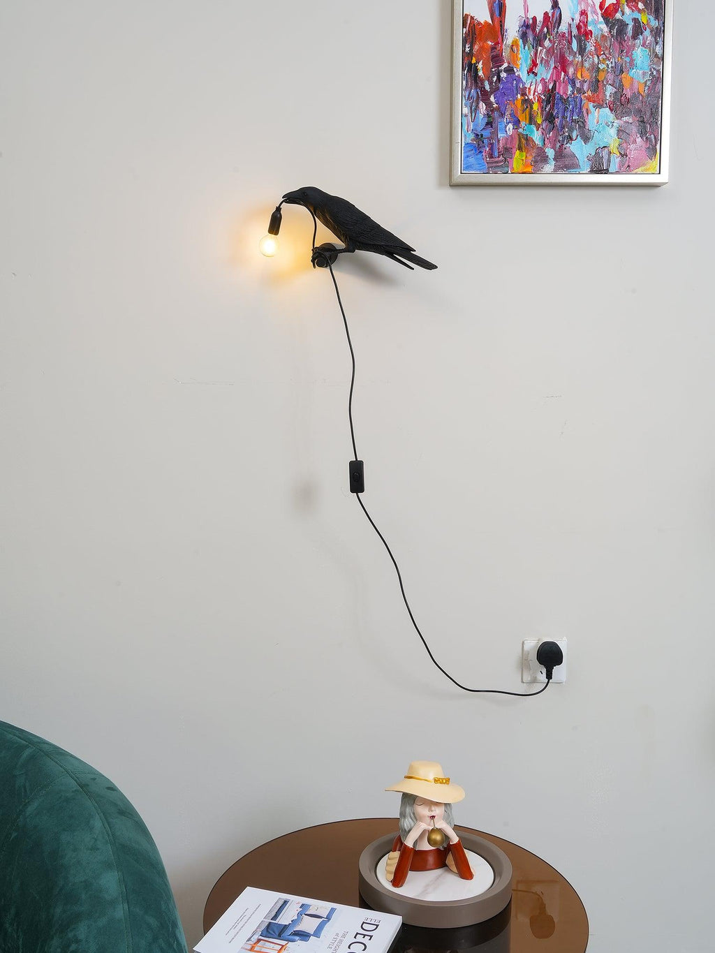 Beneath a colourful framed print, this black Clérachic bird lamp lights a round side table below with warm light.