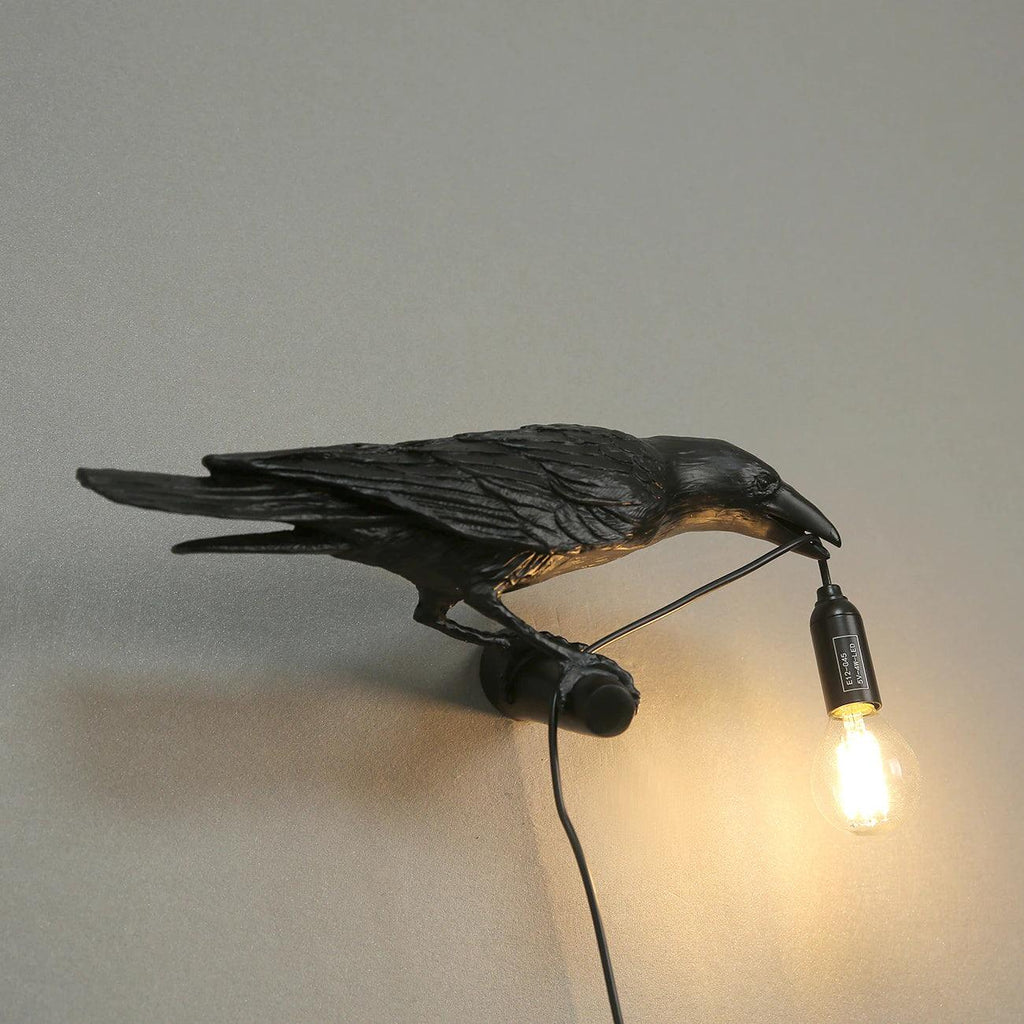 Mounted in a room corner, this black Clérachic bird lamp casts warm light down a plain beige wall.