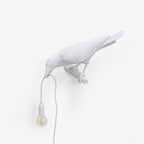 This white resin Clérachic bird lamp perches with its mount visible, beak lowered to a warmly lit hanging bulb.