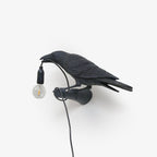 A black bird sculpture from Clérachic grips its cord in profile, the clear bulb unlit, set against a plain grey studio background.
