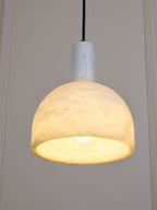 This Clérachic alabaster dome pendant hangs lit beside a panelled wall, the stone glowing amber above its black fabric cord.