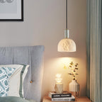Clérachic alabaster pendant light lit beside a grey tufted headboard, hanging over a nightstand stacked with books and a vase of eucalyptus.