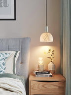 Clérachic alabaster pendant light hanging over a wooden nightstand with drawers, its warm glow lighting books and a small vase beside a grey headboard.