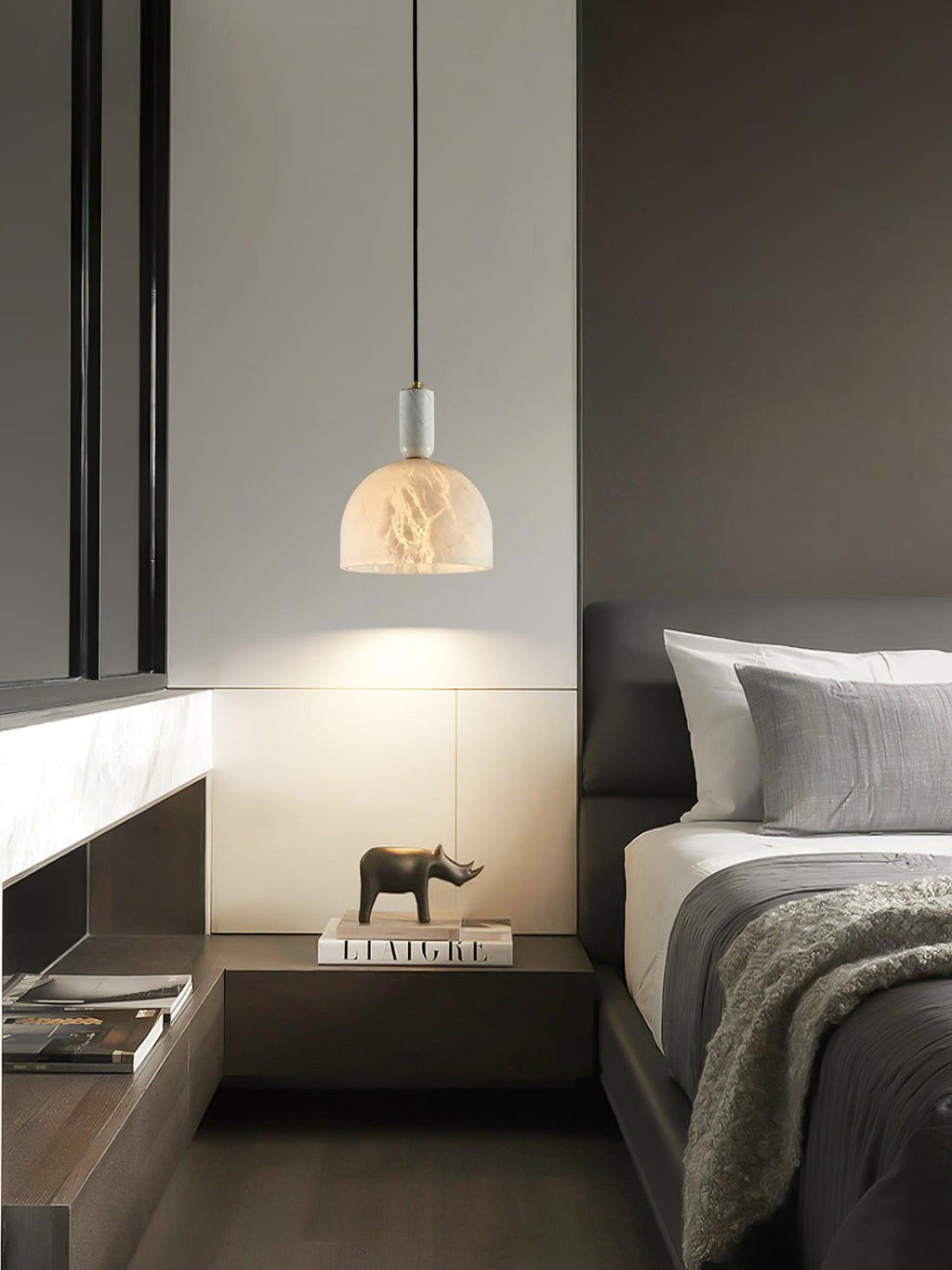 Clérachic alabaster pendant light lit above a bedside table holding a small rhino figurine and a stack of books, in a dark-wood bedroom.